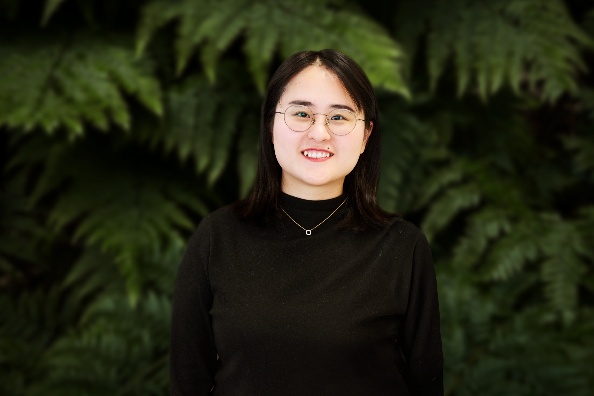 Porträt von Huifen Cong, IT Sustainability Lead bei Scania, lächelnd mit Brille und schwarzem Oberteil, vor einem Hintergrund aus grünen Farnblättern stehend.