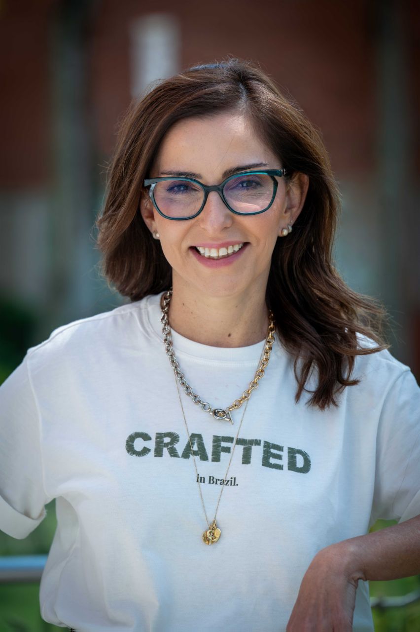 Patricia Acioli steht im Freien vor einem unscharfen Hintergrund mit Gebäuden und Vegetation. Patricia Acioli trägt eine Brille, eine helle T‑Shirt mit dem Aufdruck „CRAFTED in Brazil.“, sowie zwei Halsketten. Das Porträt ist als Nahaufnahme aufgenommen und zeigt den Oberkörper vor einem weich verschwommenen Hintergrund.