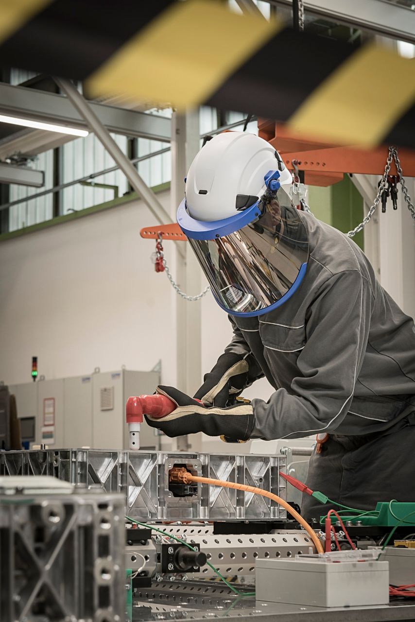 Person wearing industrial protective gear and face shield working with electrical components in a factory environment.
