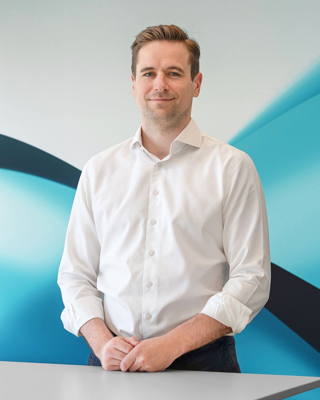 Philipp Lassernig trägt ein weißes Hemd und steht mit gefalteten Händen vor einem modernen, abstrakten TRATON-Hintergrund.