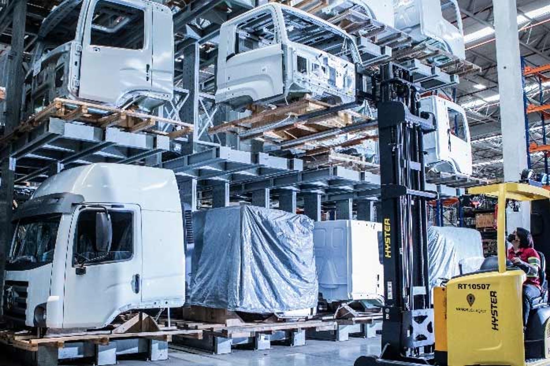 A forklift transports truck cab parts in a large industrial warehouse with stacked cabs.