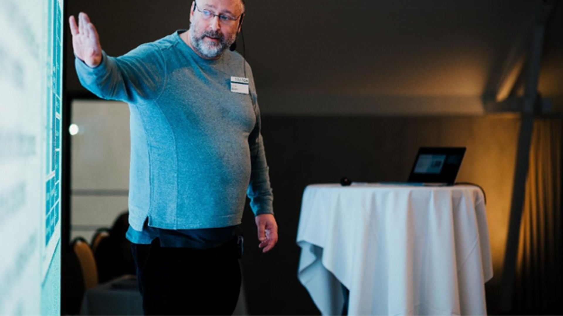 A man in a gray sweater giving a presentation in front of a screen, with a laptop and round table in the background.