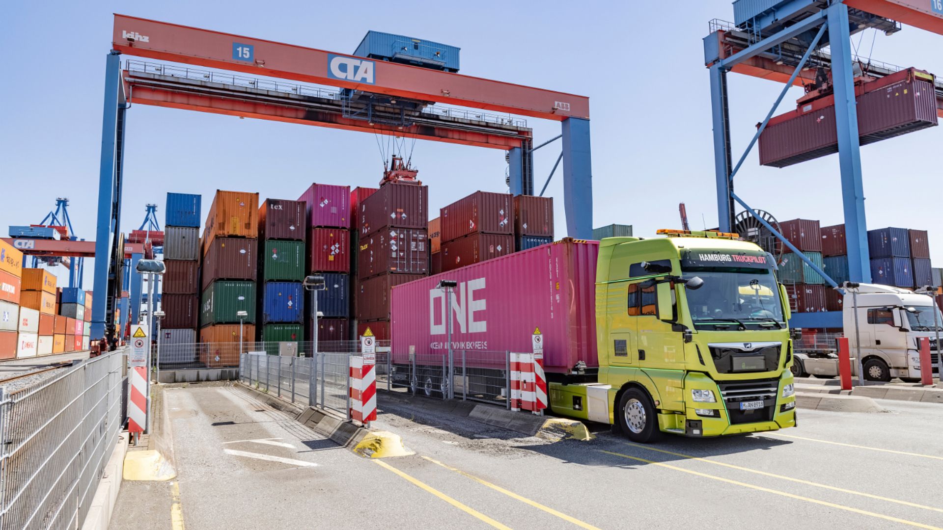 Gelber LKW transportiert einen pinken Container im Hafen, umgeben von gestapelten Containern und Kränen.