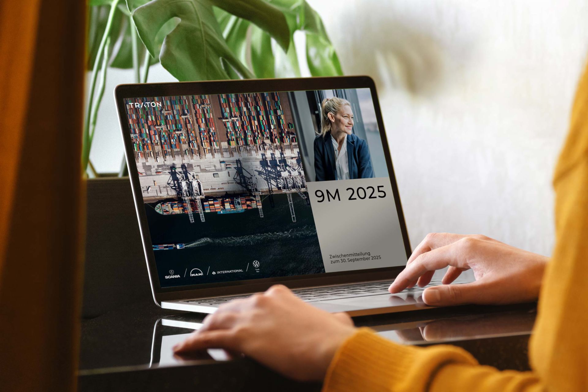 TRATON 9M 2025 Eine Person in einem gelben Pullover benutzt einen Laptop auf einem dunklen Tisch. Auf dem Bildschirm ist eine Geschäftspräsentation mit dem Titel „9M 2025 – Zwischenmitteilung zum 30. September 2025“ der TRATON Group zu sehen. Der Bildschirm zeigt ein Containerschiff aus der Vogelperspektive, ein Porträt einer lächelnden Geschäftsfrau und die Logos der TRATON-Marken, darunter Scania, MAN, International und Volkswagen Truck & Bus.