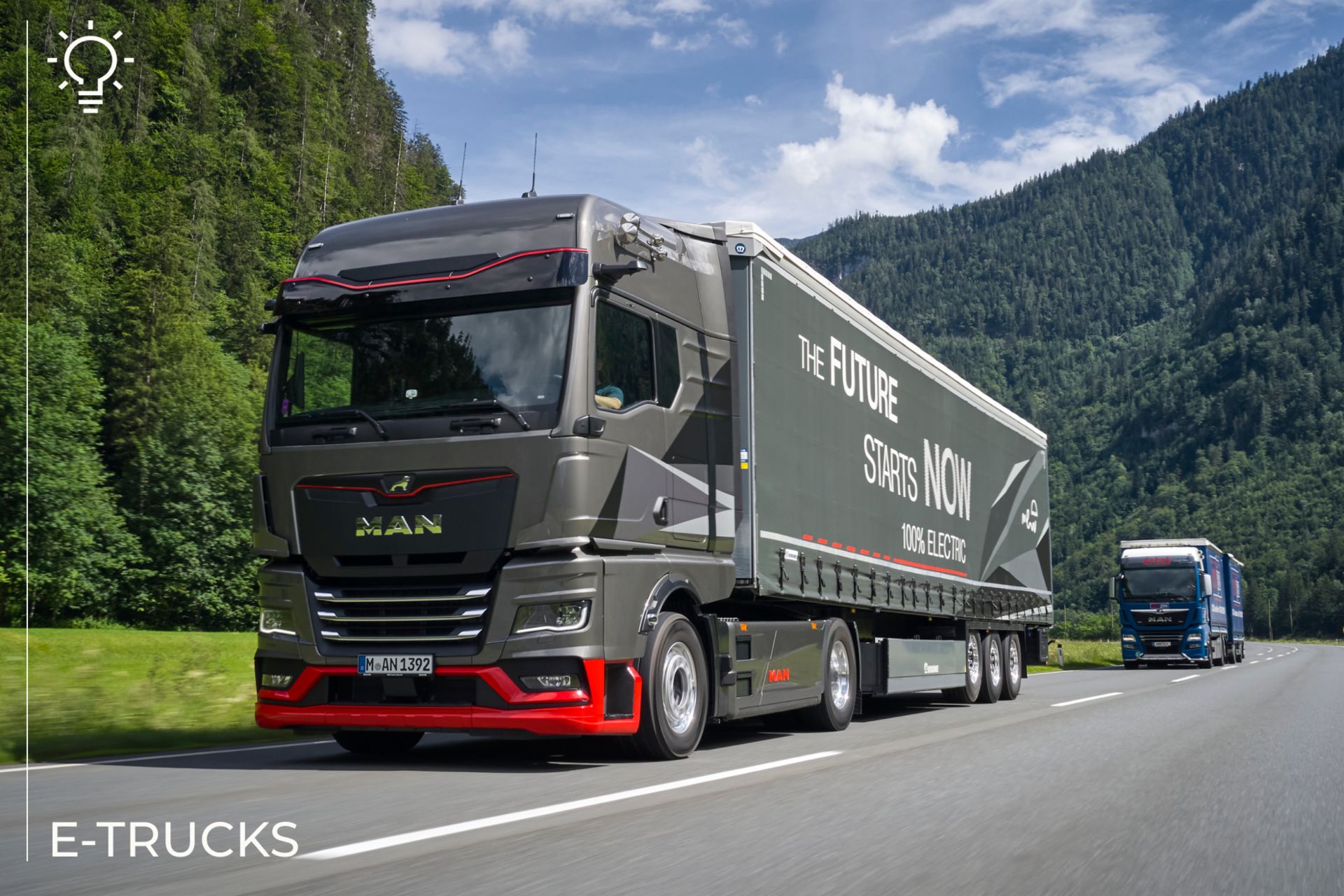 A photo of a modern electric MAN truck driving on a highway through a mountainous and forested area. The truck's trailer displays the message 