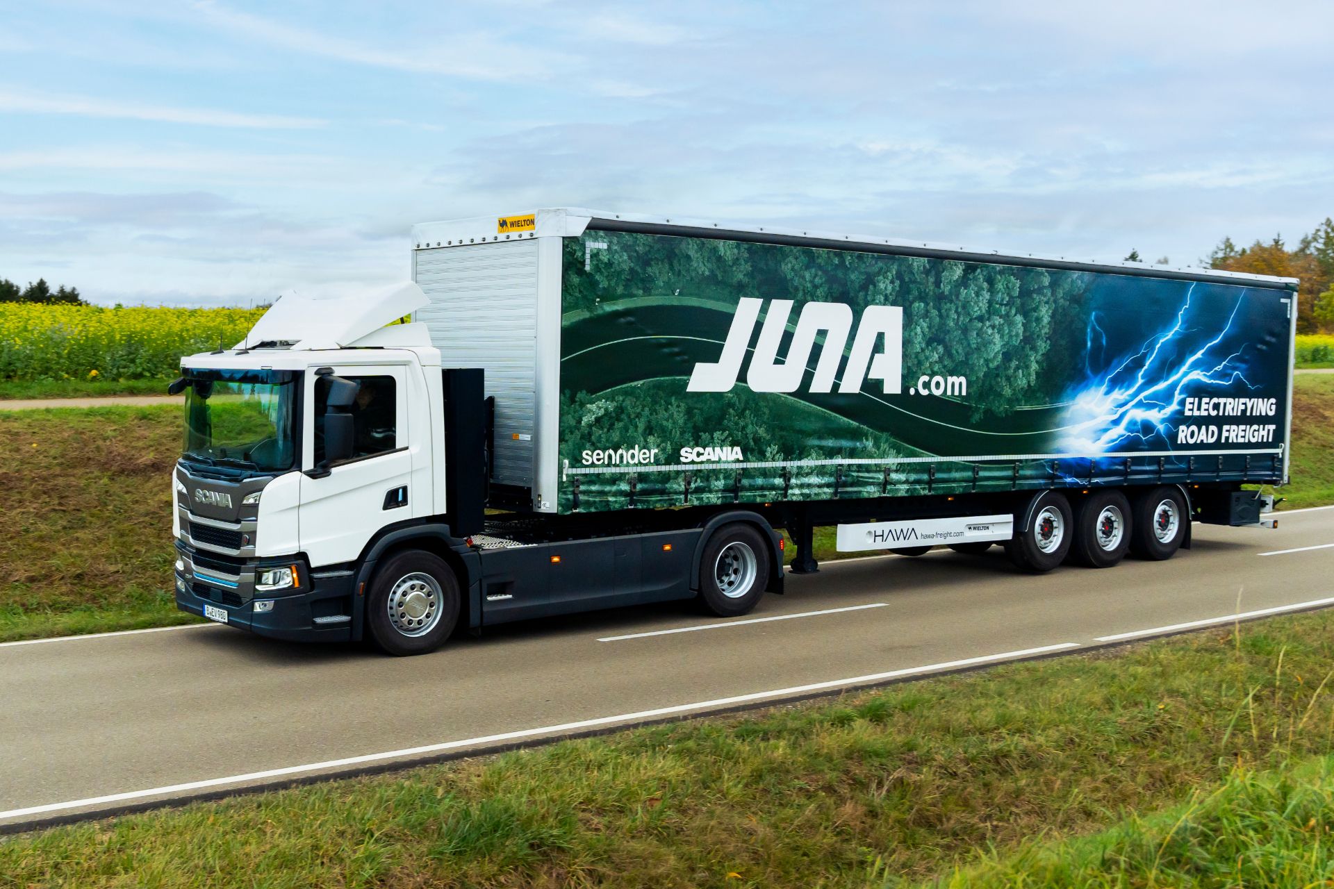 Ein elektrischer Scania-Lkw fährt durch eine ländliche Landschaft und ist Teil von JUNA, dem Joint Venture von Scania und sennder.
                 