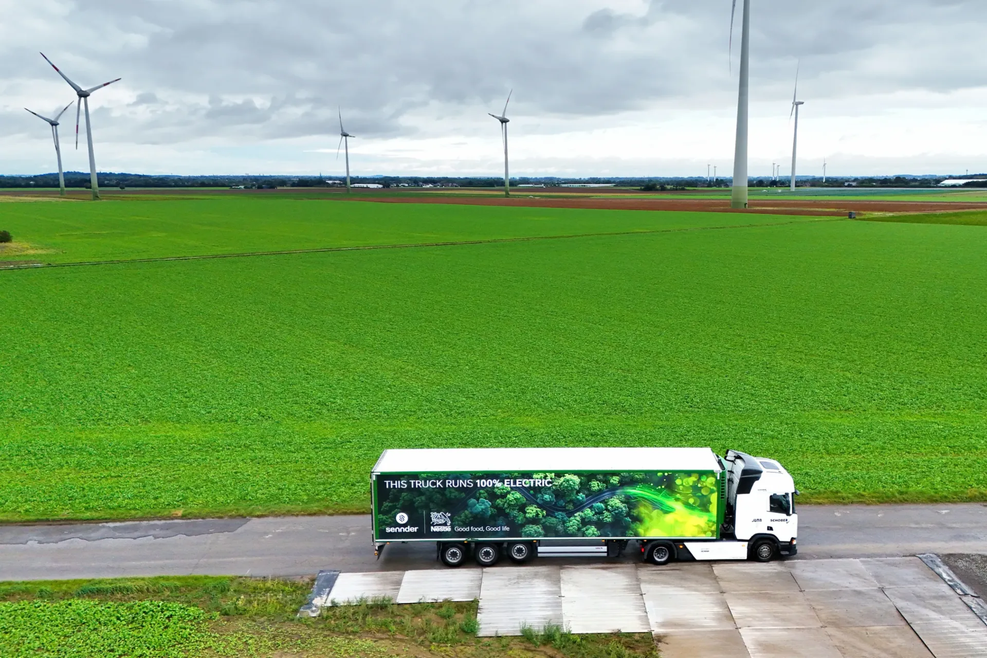 Ein elektrischer Scania-LKW fährt auf einer Landstraße durch eine grüne, ländliche Umgebung. Das Fahrzeug ist Teil des Joint Ventures JUNA von Scania und sennder, das die Elektromobilität im Straßengüterverkehr fördert.