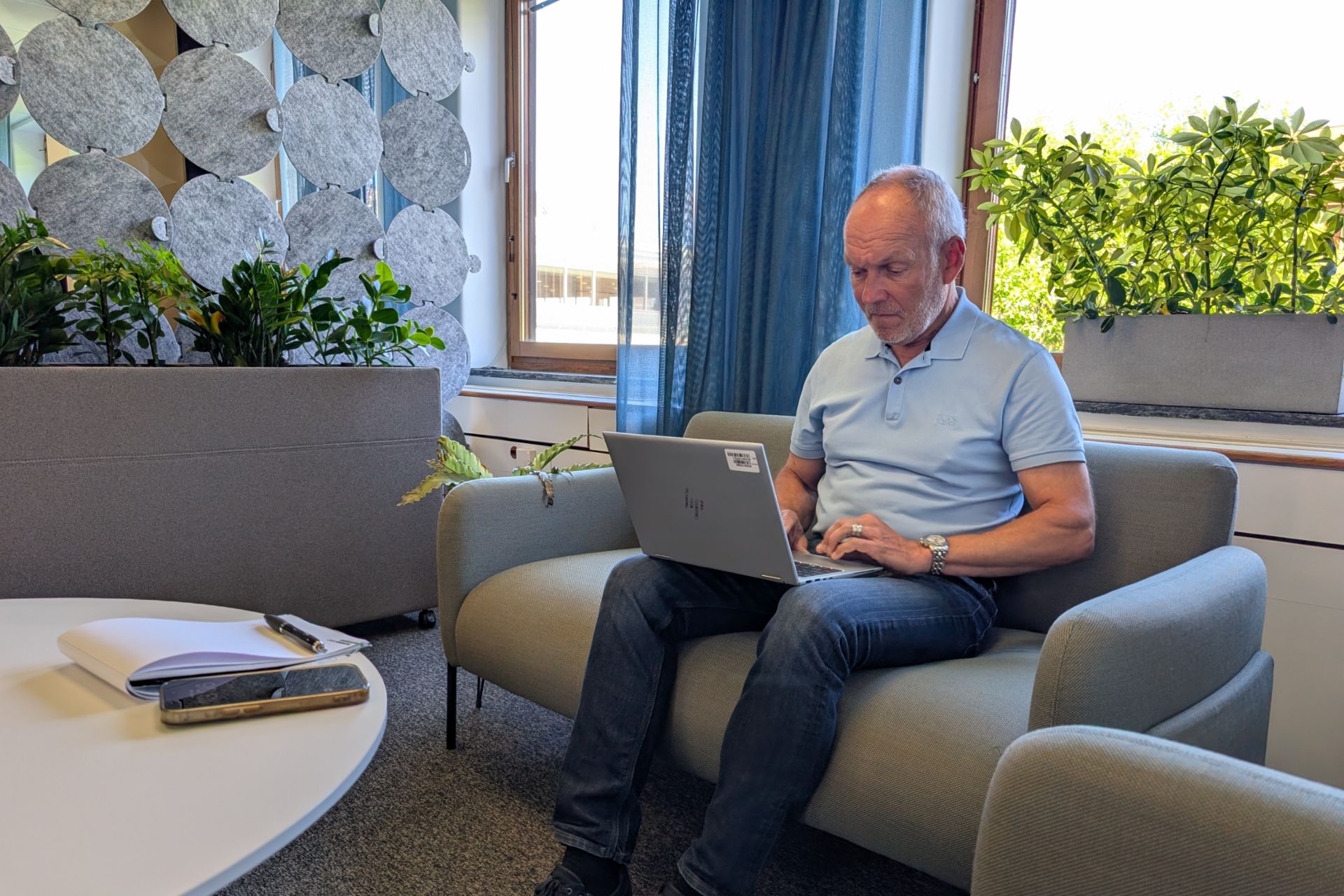  Ein Mann mit grauem Haar, bekleidet mit einem hellblauen Poloshirt und Jeans, sitzt auf einem grauen Sofa und tippt auf einem Laptop. Die Umgebung ist ein helles, modernes Büro mit Pflanzen und einer dekorativen Akustikwand aus grauen Filzkreisen. Im Vordergrund steht ein weißer Tisch, auf dem ein Notizbuch, ein Stift und ein Smartphone liegen.