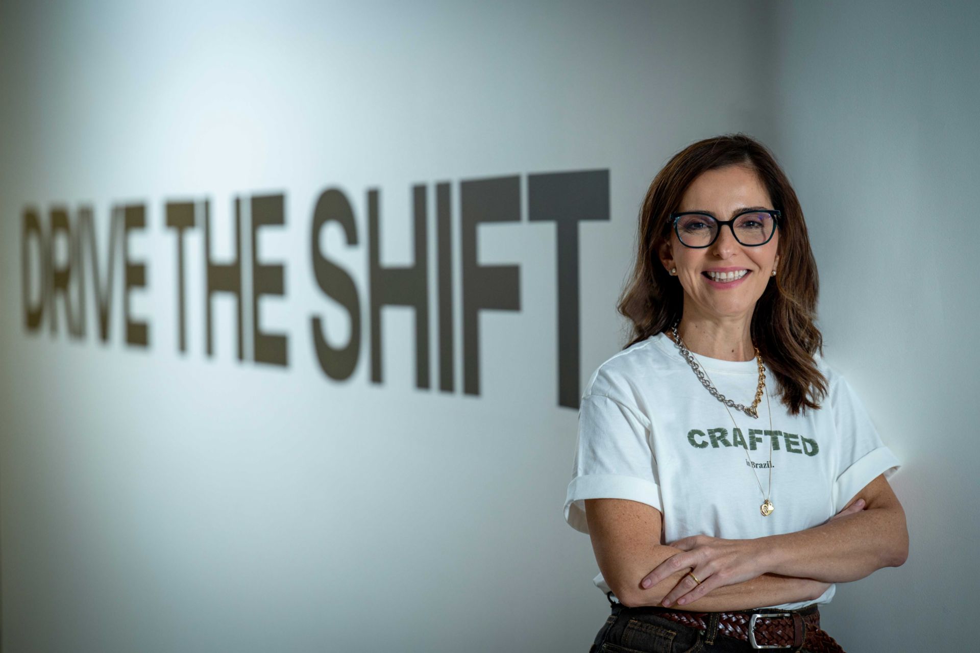 Patricia Acioli steht mit verschränkten Armen vor einer hellen Wand, auf der großformatig der Schriftzug „DRIVE THE SHIFT“ zu sehen ist. Patricia Acioli trägt eine Brille, ein helles T‑Shirt mit dem Aufdruck „CRAFTED in Brazil.“ sowie eine Halskette. Die Aufnahme zeigt Patricia Acioli vom Oberkörper aufwärts in einer Innenraumumgebung mit schlichtem Hintergrund und typografischem Designelement.