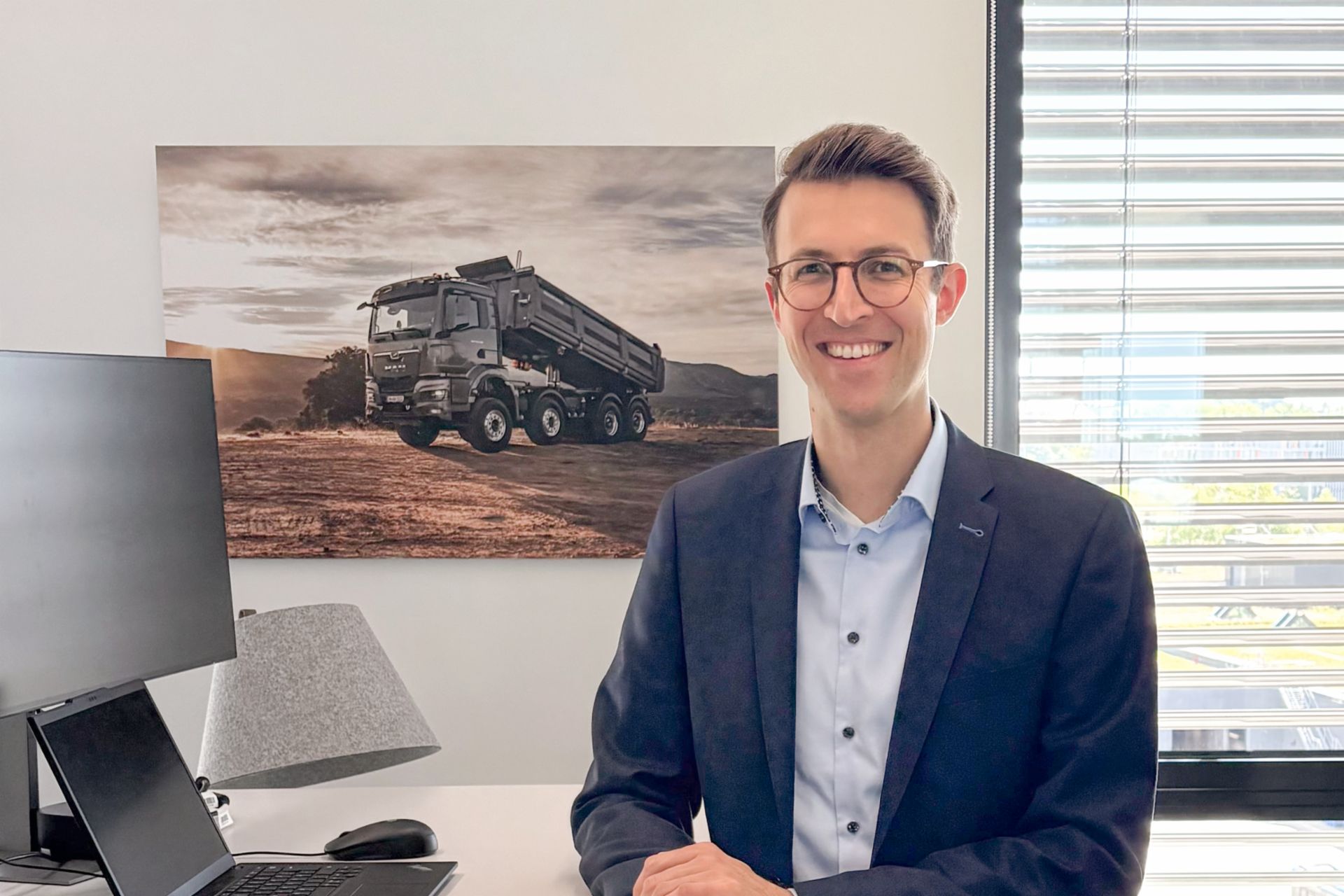 Fabio Buschle smiling in a modern office, with a picture, laptop, and window in the background.
                 