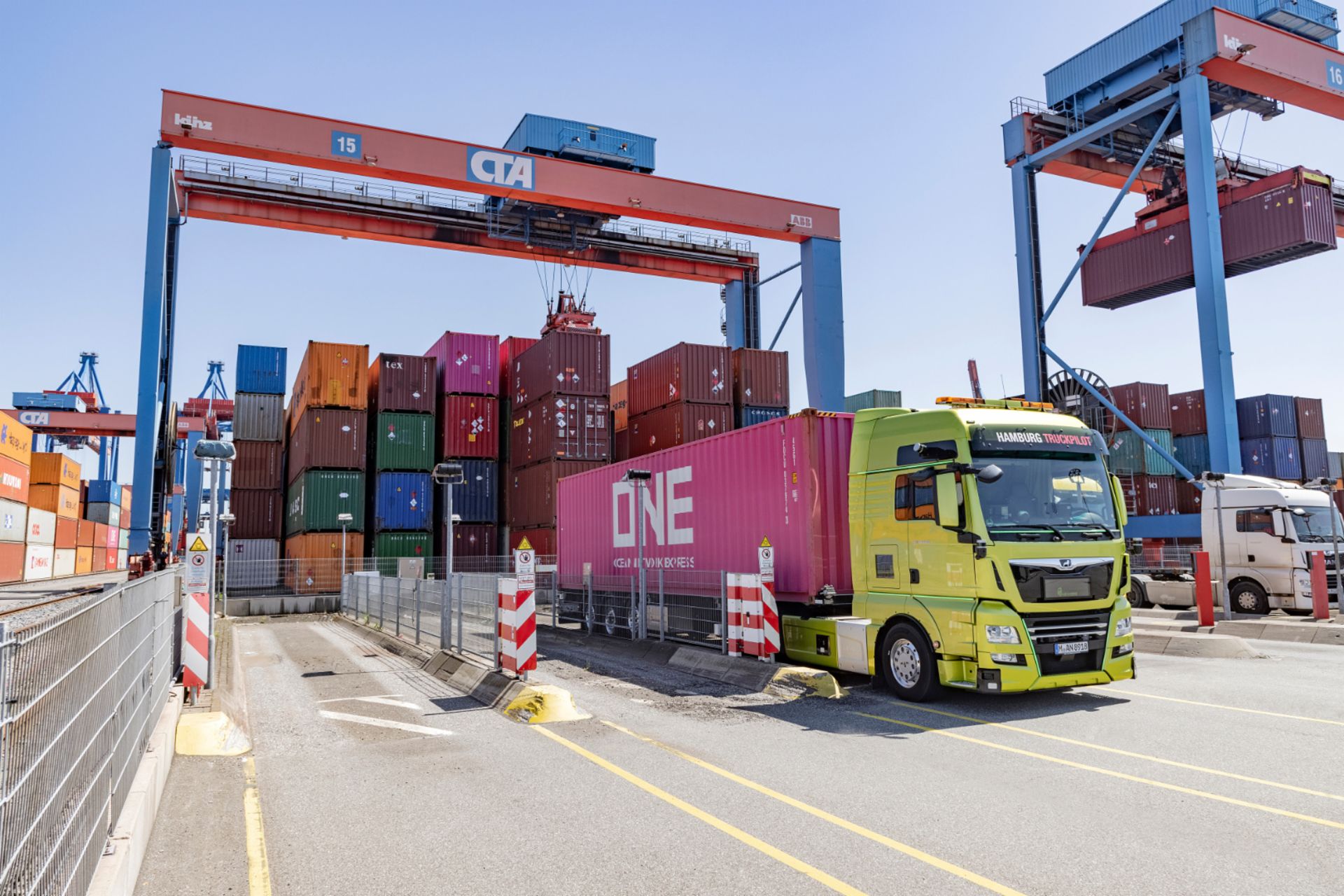 Gelber LKW transportiert einen pinken Container im Hafen, umgeben von gestapelten Containern und Kränen.